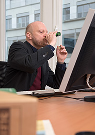 Mann am Schreibtisch trinkt hinter dem Computermonitor aus einer kleinen Flasche