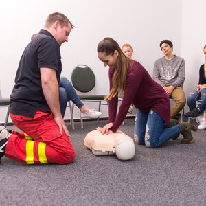 Eine Frau übt an einer Puppe die Herz-Druck-Massage, während ein Mann in Sanitätskleidung ihr gegenüber kniet.