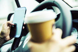 Person mit einem Becher Kaffee in der Hand bedient ein Smartphone am Lenkrad eines Autos und hält einen Kaffeebecher in der rechten Hand