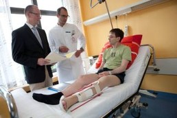 Reha-Manager der BG RCI und Arzt stehen am Bett eines am Bein verletzten Patienten und unterhalten sich mit ihm.