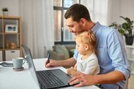 Ein Mann mit Kind auf dem Schoß arbeitet im Homeoffice am Laptop